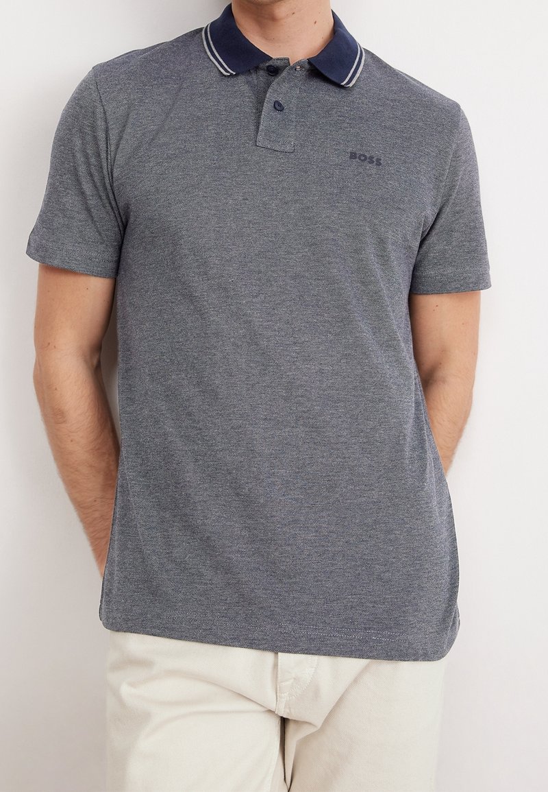 Homme portant un polo gris à manches courtes avec un col bleu marine et deux boutons, associé à un pantalon de couleur claire, sur un fond uni.