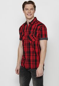 Camicia a maniche corte con bottoni in un motivo a quadri rosso e nero; presenta due tasche sul petto, dettagli neri sulle maniche e una vestibilità sagomata.