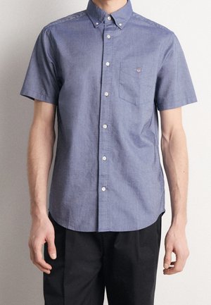 Uomo che indossa una camicia blu a maniche corte con taschino sul petto e pantaloni neri, in piedi davanti a uno sfondo semplice.