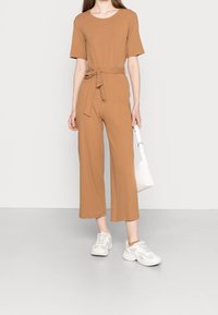 Brunrandig jumpsuit med korta ärmar, bälte i midjan och vida ben. Kombinerad med vita sneakers och en liten vit väska.