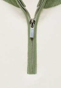 Vit plagg med en grön dragkedja och ribbade detaljer. Dragkedjan är metallisk och placerad centralt, vilket kontrasterar med tygfärgen.