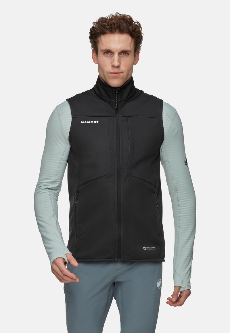 Schwarze Softshell-Weste mit Reißverschlusstaschen, hohem Kragen und Mammut-Logo. Unterlage in Hellblau mit gerippter Textur.