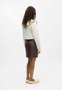 Maglione beige chiaro con colletto a volants, gonna in pelle marrone scuro e mocassini crema. Il modello è visto da dietro.