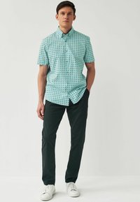 Helles blaues Kurzarmhemd mit geometrischem Muster, kombiniert mit dunklen grünen Hosen und weißen Turnschuhen. Das Hemd hat einen Kentkragen.