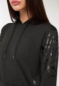 Donna che indossa un hoodie nero con toppe lucide e decorate sulle maniche e lunghe stringhe, mostrata dalla bocca fino a metà torso.