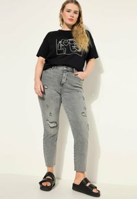 Camiseta gráfica negra con arte lineal blanco, jeans gris desgastados y sandalias negras. Diseño elegante con bolsillos y detalles de desgaste visibles.