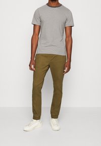 Man in een zwart-wit gestreepte T-shirt met korte mouwen, olijfgroene joggerbroeken met boorden, en witte sneakers, staande tegen een effen achtergrond.
