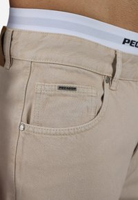 Beige denim pants with a flat front, featuring a small patch pocket and a logo tag. The waistband has white stripes and branding.