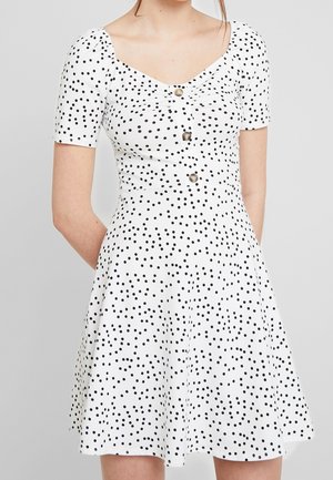 Robe blanche à manches courtes, avec un corsage ajusté et une jupe évasée, ornée de pois noirs et de trois boutons décoratifs à l'avant.