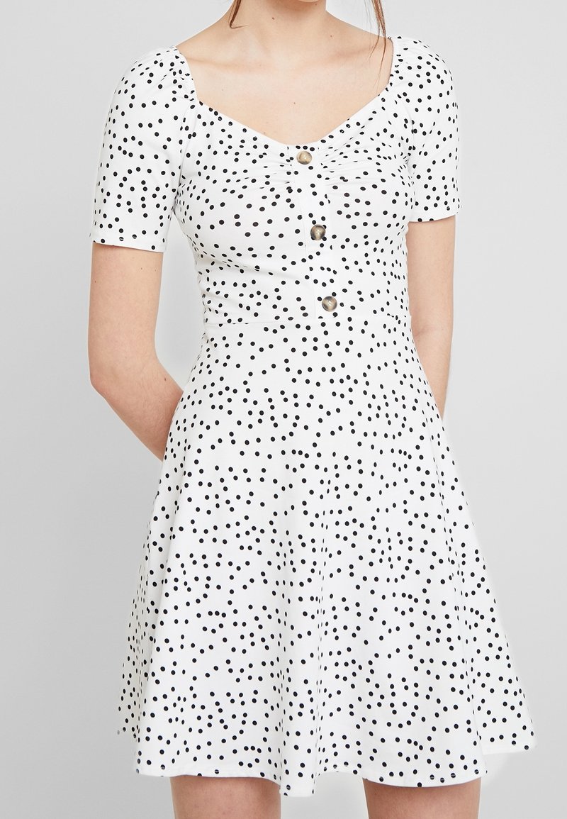 Kratší bílé šaty s krátkým rukávem, těsně padnoucím korzetem a rozšířenou sukní, zdobené černými puntíky a třemi dekoračními knoflíky na přední straně.