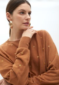 Hnědý svetr se zlatými knoflíkovými detaily, dlouhými rukávy a uvolněným střihem. Modelka má hladký účes a velké náušnice ve tvaru kruhu.