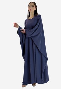 Vrouw poseert in een losse, lange marineblauwe jurk met brede, gedrapeerde mouwen en witte open teen sandalen op een lichte achtergrond.