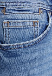 Jeans de denim azul claro com um bolso traseiro com detalhes de costura visíveis. Inclui acessórios em metal e um acabamento texturizado.