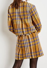 Conjunto de chaqueta y falda a cuadros en amarillo, morado y blanco, confeccionado en tela texturizada. Presenta un dobladillo sin rematar y un patrón clásico de cuadros.