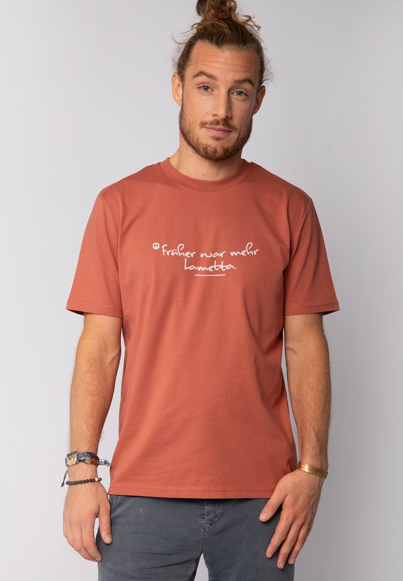 Man met opgestoken haar, gekleed in een roestkleurig T-shirt met witte Duitse tekst, grijze broek en verschillende armbanden, staande tegen een effen achtergrond.