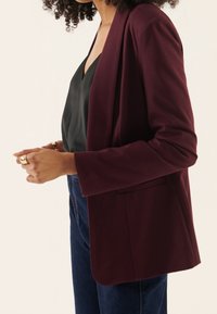 Femme portant un blazer bordeaux foncé, un haut noir à col en V et un jean bleu, vue de profil à hauteur de la taille, mains proches l'une de l'autre.