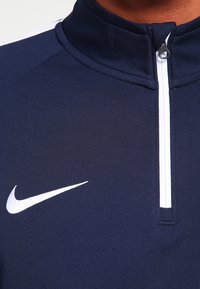 Marint träningsjacka med hög krage, tillverkad av textil med struktur. Har en vit dragkedja och en Nike-logotyp med swoosh.