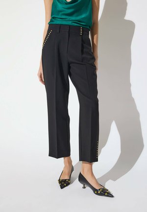 Mujer con pantalones cortos negros con tachuelas doradas, una blusa verde de satén y zapatos de tacón bajos puntiagudos negros con tachuelas doradas.