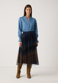 Denimbluse mit knöpfbarem Vorderteil, langen Ärmeln und Rüschenkragen, kombiniert mit einem geschichteten Tüllrock in Marine- und Brauntönen, getragen mit knielangen Stiefeln.