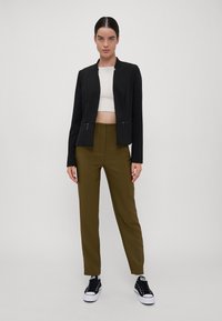 Black cropped blazer with zip pockets, paired with olive green high-waisted pants and black sneakers. Smooth fabric and tailored fit.