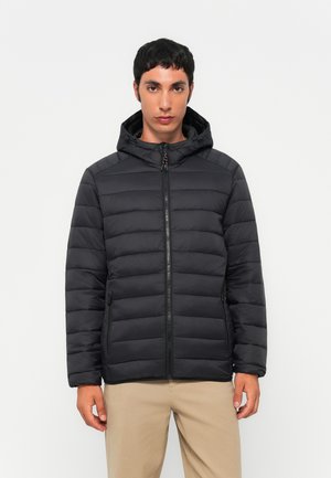 Mand iført en sort quiltet hættetrøje med lynlås, stående mod en ensfarvet lys baggrund, med beige bukser.