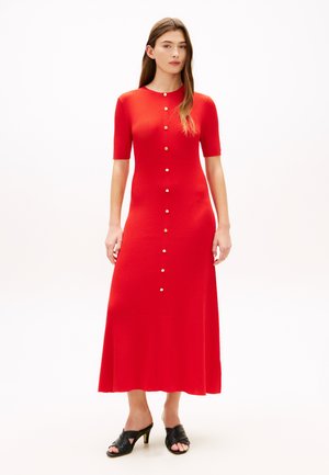 Kvinne stående iført en rød ribbestrikket midikjole med gullknapper foran og svarte heeled mules mot en hvit bakgrunn.