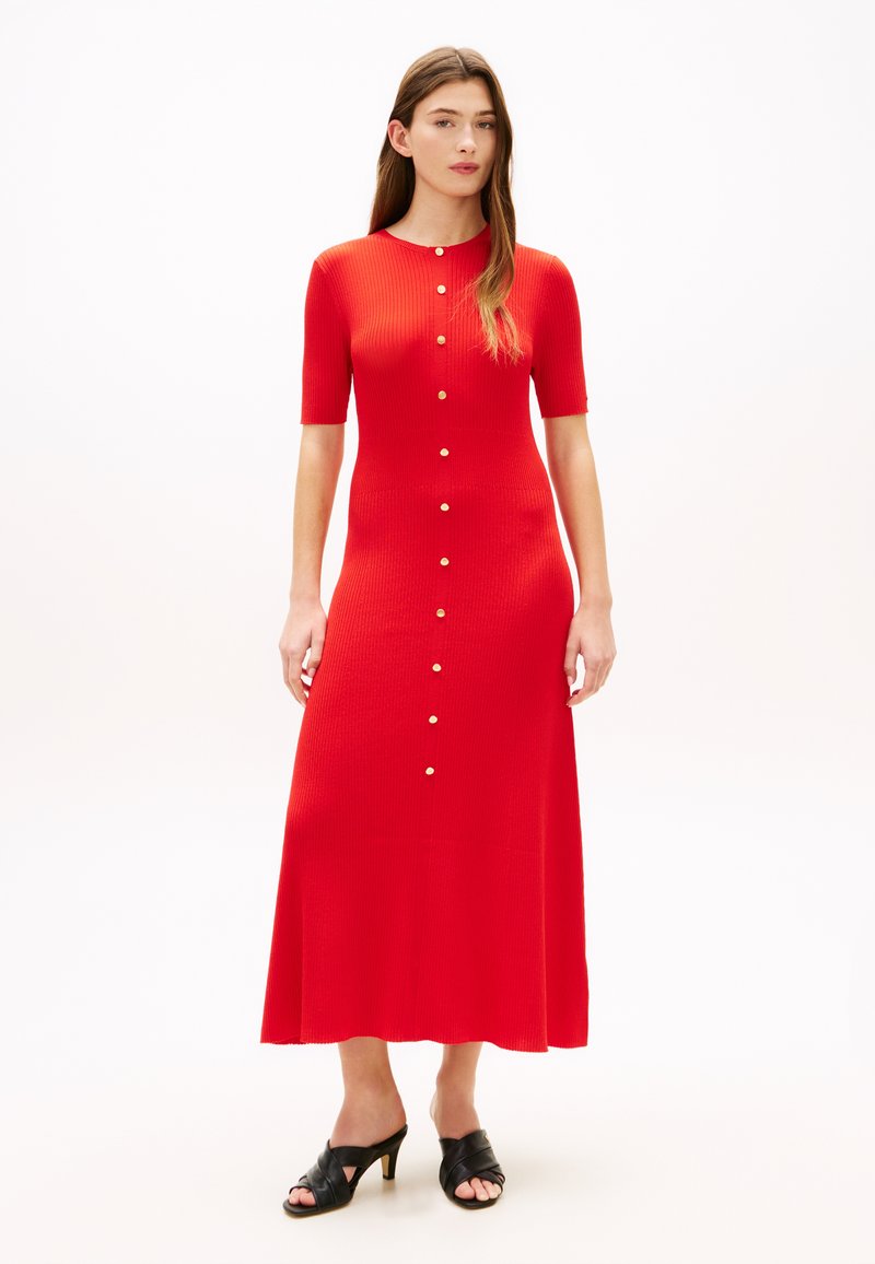 Γυναίκα που φοράει κόκκινο ριμπ midi φόρεμα με χρυσά κουμπιά μπροστά και μαύρα mules με τακούνι, στέκεται μπροστά σε λευκό φόντο.