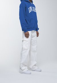 Felpa blu con scritte bianche, pantaloni cargo bianchi con tasche e sneakers bianche. Il tessuto appare morbido e dallo stile informale.