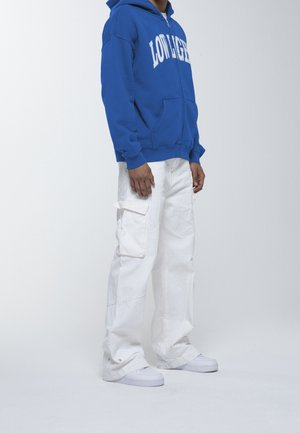 Felpa blu con scritte bianche, pantaloni cargo bianchi con tasche e sneakers bianche. Il tessuto appare morbido e dallo stile informale.