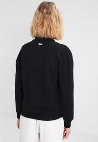 Persona con cabello rubio vistiendo un suéter negro de FILA y pantalones blanco a rayas, mostrada desde atrás contra un fondo sencillo.