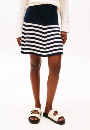 Femme portant une jupe rayée bleu marine et blanc, un pull en maille crème et des sandales blanches avec des boucles dorées, debout sur un fond blanc.