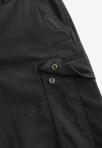 Schwarzes Stoffkleidungsstück mit einer gefalteten Tasche mit Druckknopfverschluss, sichtbaren Nähten und einer strukturierten Oberfläche.
