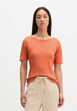 Jeune femme aux longs cheveux foncés, portant un t-shirt orange à manches courtes et un pantalon beige à cordon, debout devant un fond blanc.