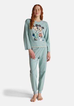 Jonge vrouw met rood haar, gekleed in lichtblauwe Mickey en Minnie Mouse pyjama's, staand op blote voeten tegen een witte achtergrond.