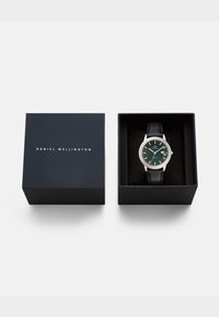 Daniel Wellington pulksteņa iezīmes ir zaļa ciparnīca, sudraba korpuss un melns ādas josta, kas ievietota tumši zilā kastē ar zelta burtiem.