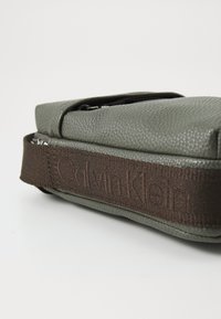 Sivá textúrovaná taška s hnedým látkovým popruhom. Popruh má vo vzore vyrazené logo "Calvin Klein". Uzavretie na zips.