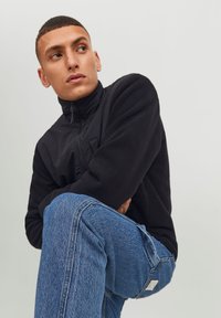 Svart fleece-jakke med høy krage, glidelås foran og brystlomme, sammen med blå denim jeans som har klassisk passform og tekstur.