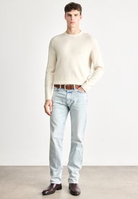 Gaiši bēša, apaļās apkakles džemperis, kombinēts ar izbalējušām zilām džinsu biksēm un tumši brūnām ādas potīšu zābakām. Vienkāršs un piegulošs dizains.