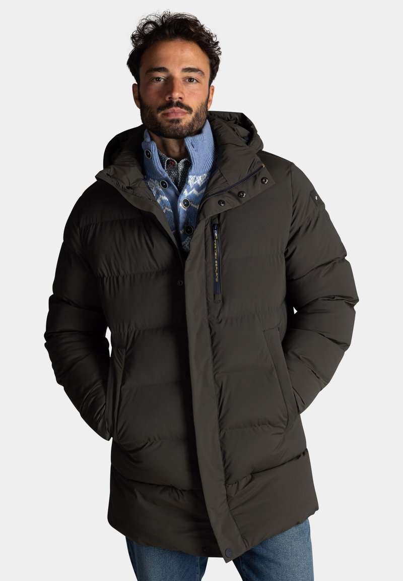 Donkergroene pufferjas met een capuchon, een ritssluiting aan de voorkant, knoopaccenten en horizontale quilt. Gecombineerd met een blauwe geblokte trui daaronder.