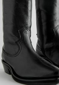 Bottes de cowboy en cuir noir avec un bout pointu, des accents de couture et une fermeture éclair sur le côté ; texture lisse et talon empilé.