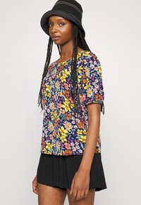 Top de manga curta floral com um fundo azul-marinho e flores multicoloridas. Combinado com uma saia preta plissada e um chapéu de balde preto com riscas.