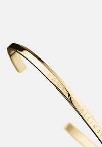 Gold-tone metal cuff bracelet with a smooth, polished surface and a subtle engraving of "DANIEL WELLINGTON." Slim, curved design.