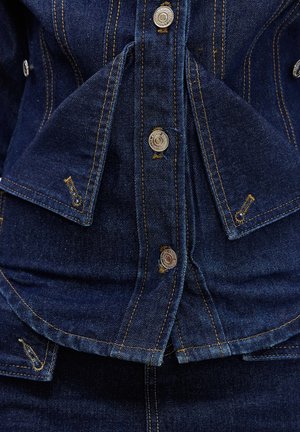 Veste en denim bleu foncé avec boutons-pression argentés et coutures contrastantes jaunes sur les rabats pliés et les coutures.