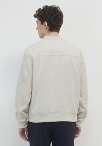 Giacca bomber di colore beige chiaro con texture liscia, maniche a costine e vita a coste. Caratterizzata da un design semplice e colletto rotondo, mostrata da dietro.