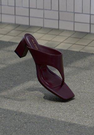 Dark red high-heeled sandal with a square toe and wide strap, positioned on a tiled and asphalt surface background.