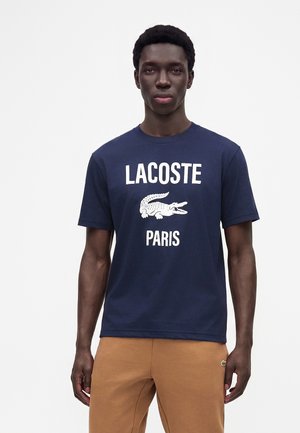 Jeune homme portant un t-shirt Lacoste Paris bleu marine et un pantalon marron clair, debout devant un fond blanc uni.
