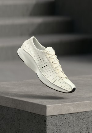 Λευκό διάτρητο αθλητικό παπούτσι Nike με κορδόνια, αιωρούμενο πάνω από μια τσιμεντένια άκρη, με θολό γκρι σκηνικό σκάλας στο φόντο.