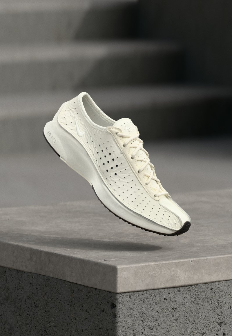 Λευκό διάτρητο αθλητικό παπούτσι Nike με κορδόνια, αιωρούμενο πάνω από μια τσιμεντένια άκρη, με θολό γκρι σκηνικό σκάλας στο φόντο.