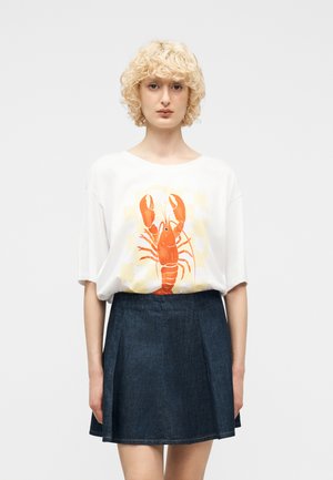 Osoba z kręconymi blond włosami, ubrana w białą, oversize'ową koszulkę z dużym pomarańczowym nadrukiem homara i ciemną spódnicę dżinsową, stoi na tle białego tła.