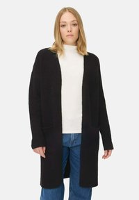 Langer schwarzer Cardigan aus einem weichen, strukturierten Stoff. Mit offenem Vorderteil, langen Ärmeln und zwei vorderen Taschen. Über einem weißen Oberteil getragen.